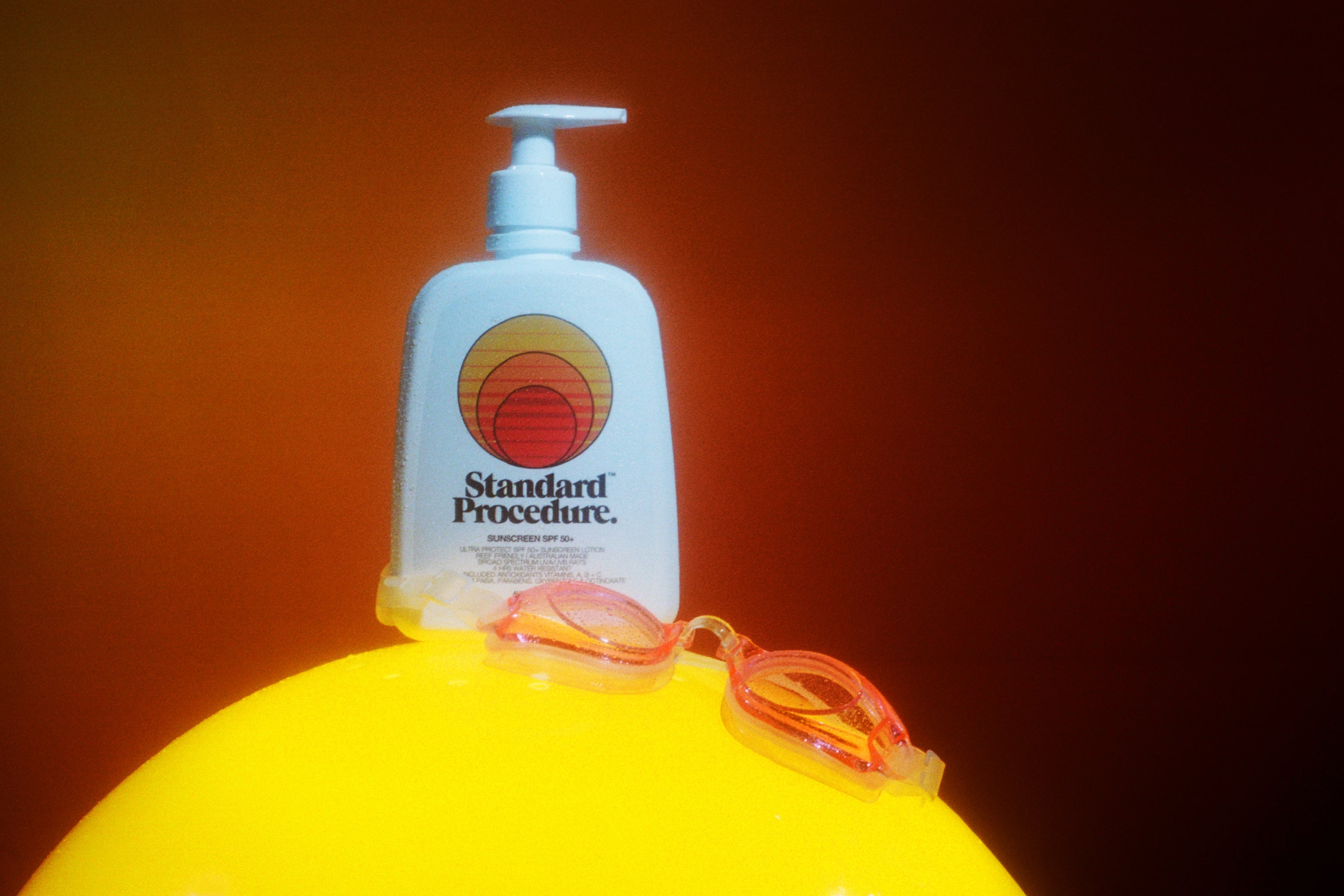 Sunblock lotion SPF 50 bottle placed on a bright yellow surface with swimming goggles against a warm orange background.