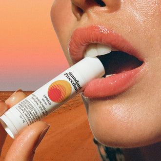 Lip protection SPF 50 balm held near glossy lips with a desert background and warm sunset lighting.