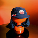 Blue surf hat displayed on an orange stand with a wide brim, neck flap, and adjustable chin strap, featuring the Standard Procedure logo for sun-safe beachwear